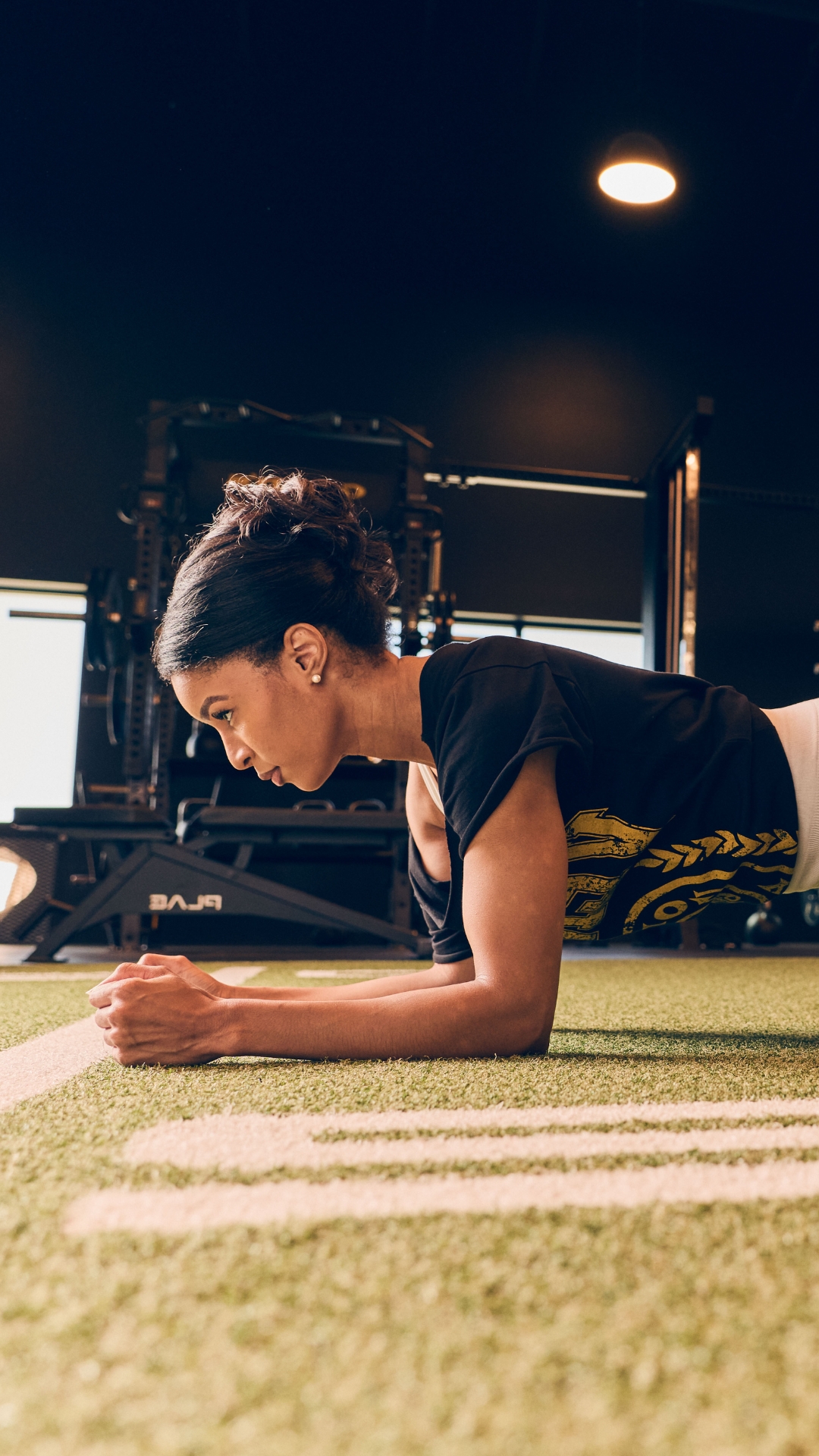 Woman holding a strong plank on indoor turf at Gold’s Gym Greeley with racks and strength equipment in the background in a serious training environment